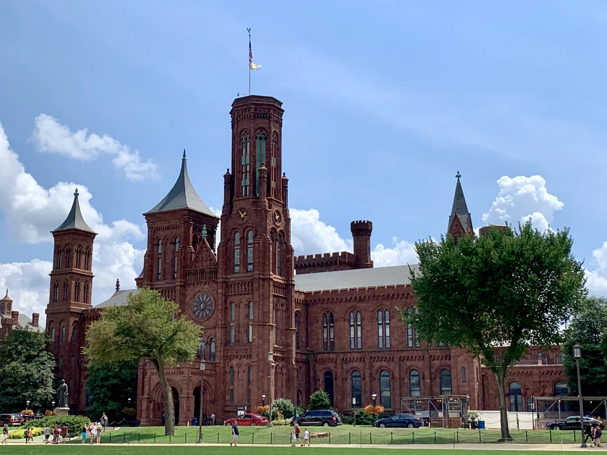 Red sandstone Smithsonian Castle with towers, lawn, trees and visitors under a blue sky.
