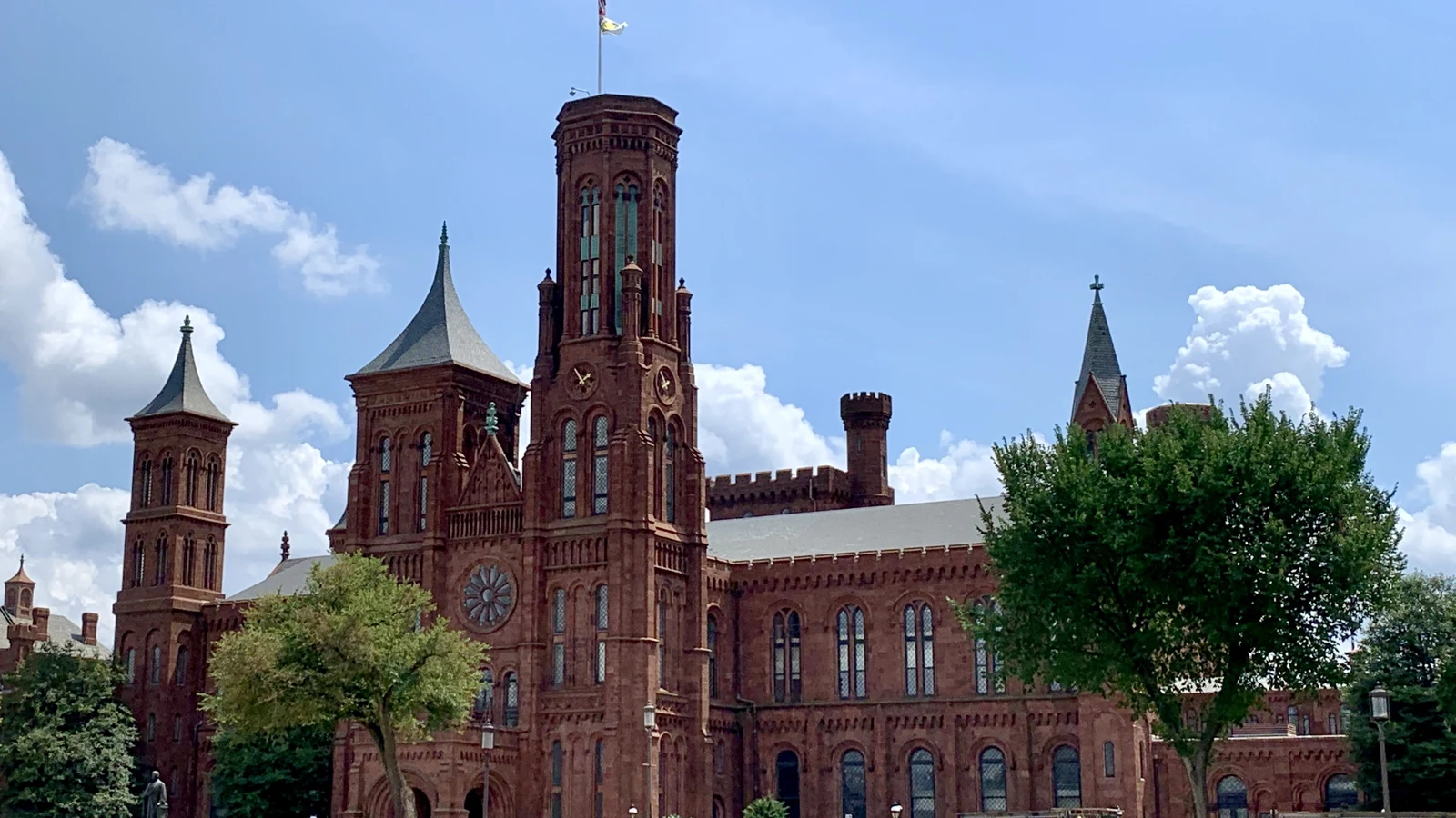 Red sandstone Smithsonian Castle with towers, lawn, trees and visitors under a blue sky.