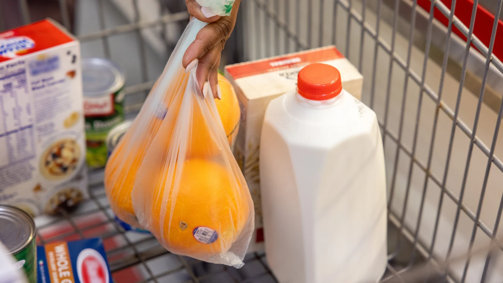 A shopper places a bag of oranges into a grocery cart beside milk and packaged food.