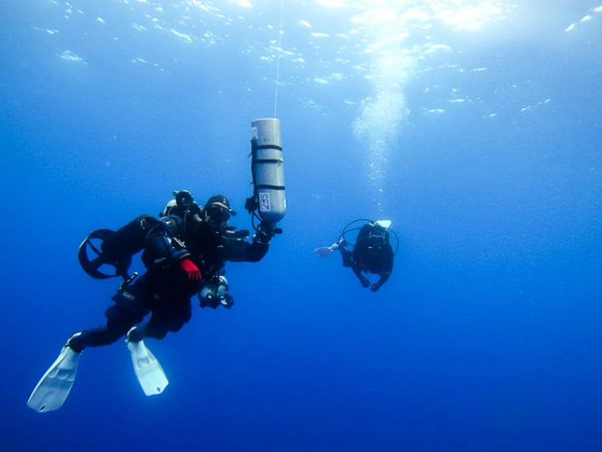 Two scuba divers pause in blue water, one wearing a closed-circuit rebreather and the other conventional scuba equipment.
